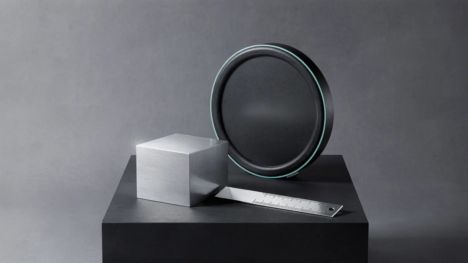 A minimal graphic still life on a plain dark plinth: a smooth brushed-steel block with an unmarked face, a slender polished measuring rule blank along its length, and behind them a dark round instrument dial with an empty face and no needle, against a seamless graphite background.
